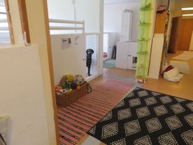 Rum med ljust golv, mattor i olika färger, en korg med hundleksaker, en svart hund som kikar fram bakom en gallergrind